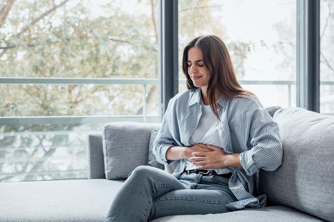 Woman sitting on a sofa holding her lower abdomen, representing pelvic pain and discomfort associated with uterine fibroid symptoms.